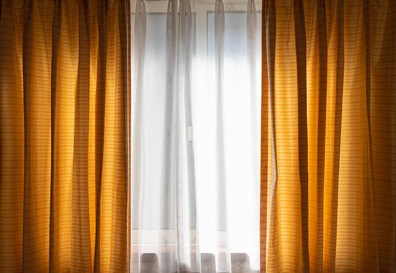 A bright window framed by golden-yellow drapes, with sheer white curtains softening the light in the middle.