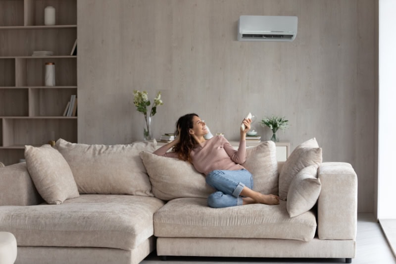 Relaxed female homeowner sitting on a comfortable couch, turning on the ductless unit with a remote